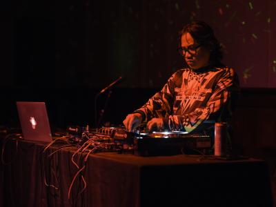 Wok the Rock stands at a DJ table, adjusting the dials on his electronic equipment. He wears black glasses, and a patterned long-sleeved shirt. He’s illuminated by a red light, and green and red lasers pattern the wall behind him.