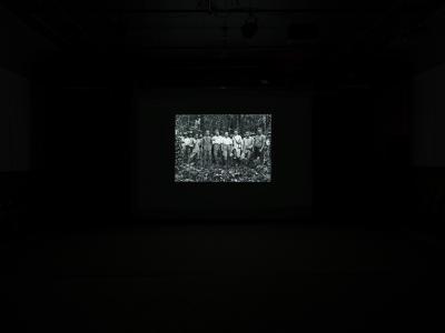 A still from a film projected in the Grand Luxe Hall that shows an archival group photo. Set in a forest, the black-and-white image depicts six white men standing in front of a group of Indonesian men.