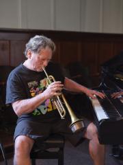Bill Clark blows into his trumpet while playing piano with his left hand. He is seated at a piano bench and wears a black Vancouver International Jazz Festival T-shirt and black shorts.