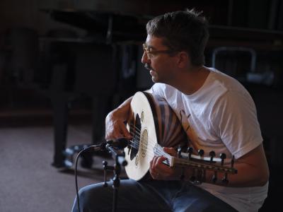 Aram Bajakian sits on a piano bench in the Grand Luxe Hall and plays an oud. A microphone points to the body of the instrument. Aram wears a white graphic T-shirt, blue jeans, and round glasses.