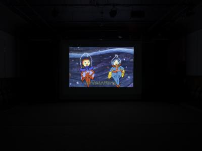 A still from a film projected in the Grand Luxe Hall. Two cartoon space creatures, one red and one blue, float in outer space. A caption reading, They replicate in an infinite number to form the universe we live in today, is written across the bottom of the image in a squiggly yellow font.
