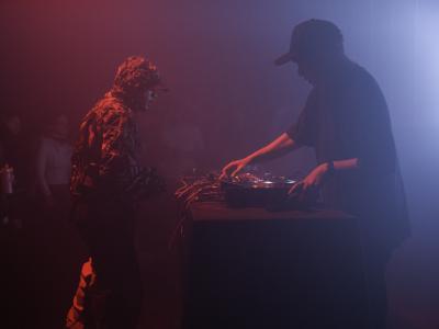 Kasimyn stands at a DJ booth and turns dials on a CDJ. He wears a black baseball cap and black t-shirt. Ican Harem faces him, and wears a hooded jacket and baseball cap. Smoke from a fog machine diffuses the light in the room into a soft red and blue glow.