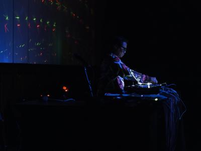 Wok the Rock stands at a DJ table in a dark room. He is illuminated by the light emitted from his DJ equipment, and green and red lasers are projected on the back wall. 