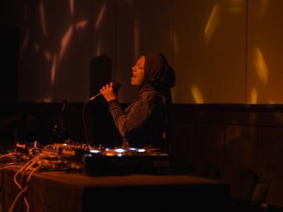 Lit by a warm red light, Rani Jambak stands behind a DJ table and sings into a microphone with her eyes closed and head thrown back. She wears headphones over her head scarf, and is dressed in a sparkly top.
