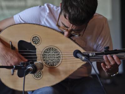Seated, Aram Bajakian gazes down while playing oud. Two microphones point to the body of the instrument. He wears round amber glasses, a white T-shirt, and blue jeans.