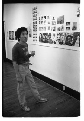 A person with a shag-style hair cut, and long sleeve shirt rolled up to their elbows, addresses the camera and holds a glass goblet in their left hand. In front of them are a series of portrait photographs that are pinned-up to a gallery wall. 
