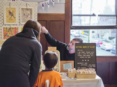 A man wearing a black hoodie sits at a table and points at a graphite drawing of a cat clipped on a pegboard. A woman in a black jacket and a child dressed in an orange shirt with a white number one on the back face him. The table is decorated with wooden boxes holding hand drawn buttons, and a sign that reads: Buttons made by Elliot. Hello, I am nine years old and I love to draw, paint, and make all sorts of art. Magnets and buttons 4 dollars each, 4 for 15 dollars. Art Prints 20 dollars.