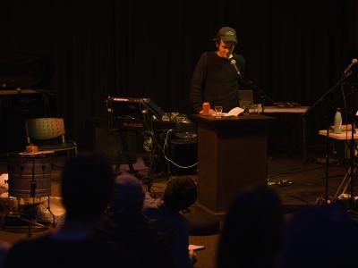 Hamish Ballantyne reads at lectern. Speaking into a microphone, he gazes down at his book positioned next to a can of beer and a glass of water. He is illuminated by an overhead orange light, and wears a baseball cap and long sleeved shirt. Various instruments, cables, and microphones are arranged behind him. 