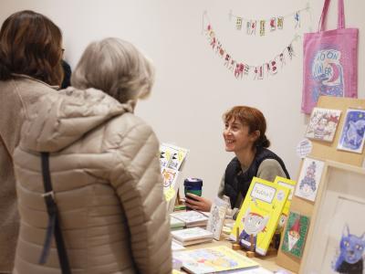 Lisa Cinar sits behind a table with a display of her books, prints, and illustrations. A pink illustrated tote bag and a pennant banner made from small drawings are hung on the wall behind her. Lisa holds a green reusable coffee cup and smiles at two customers dressed in beige coats.