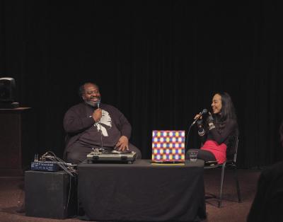 Darius Jones and Harmony Holiday are seated at a table in the Grand Luxe Hall. They each hold microphones, and share a laugh while in conversation. A turntable, a copy of Darius’s LP, and a glass of water are positioned on the table in front of them. Darius wears glasses and a black cardigan over a black T-shirt with a portrait of Sade, and Harmony wears black fingerless gloves, a black turtleneck, and a hot pink mini skirt over black ribbed tights.