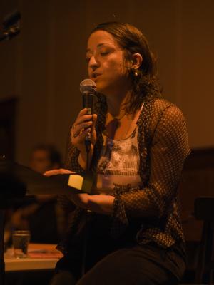 Tiziana La Melia holds a microphone in her right hand as she reads from material propped on a music stand. She wears a sheer patterned shirt over a white ribbed tank top. She’s illuminated by an orange light. 