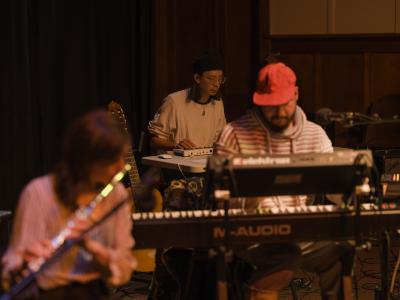 Julian Hou is seated and plays synthesizer. He wears glasses, a black toque, and a jade pendant over a white long sleeved shirt that is bunched at the elbows. Captured with a shallow depth of field, Prince Nifty and Amy Gottung can be seen playing keyboard and flute in the foreground of the image.