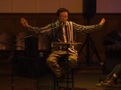 Basked in an orange glow, Fan Wu sits on a tall wooden chair and gazes down at material on a music stand. His arms are energetically outstretched as he reads into a microphone. He wears glasses and a beaded necklace over a striped collared shirt. 
