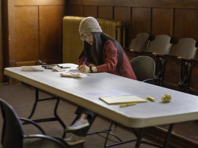 Josephine Lee writes at a grey folding table in the Grand Luxe Hall. She wears a beige knit toque, glasses, and a red sweater.