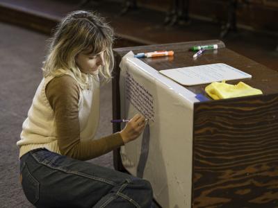 Lauren Lavery writes on an erasable surface taped to a wooden lectern laid on its side. She sits on the floor, and wears a beige sweater vest over a brown long sleeve shirt, and blue jeans.