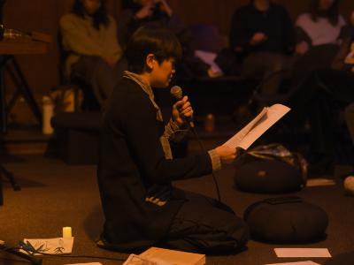 Seen in profile, S F Ho kneels on the floor of the Grand Luxe Hall. They hold loose papers in their left hand and are reading into a microphone held in their right hand. Other papers and books, as well as a small candle and meditation pillows, are arranged on the floor around them. They are bathed in warm orange light, and wear a plaid shirt, a black jacket, and black pants with frog closures down the leg.