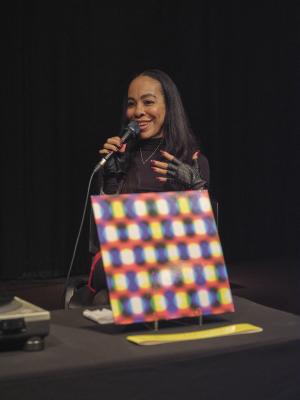 Harmony Holiday smiles into a microphone held in her right hand. Her left hand is placed on her chest, and is accessorized with black leather studded fingerless gloves. She is seated at a table covered in a black table cloth where an LP is propped up. The album cover is a pattern of white, blue, and yellow overlapping circles on a red background.