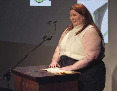 Samantha Marie Nock stands at a lectern in the Grand Luxe Hall and smiles toward the audience. She is dressed in a white sleeveless turtleneck, and has tattoos on her forearms. Her book is set down on the lectern next to a can of Montellier soda. 