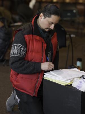 Emiliano Sepulveda kneels on the floor of the Grand Luxe Hall while using a black wooden box as a writing surface. He wears his hair in a braid, and is dressed in a red puffer vest layered over a knit sweater.