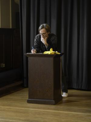 Christian Newby writes on a whiteboard placed on a wooden lectern. He leans on the surface with his chin resting in his left hand.