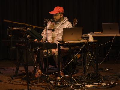 Prince Nifty is seated between a synthesizer, and a table holding a laptop and various electronic instruments. He sings into a microphone and wears a pink baseball cap and striped hoodie. 