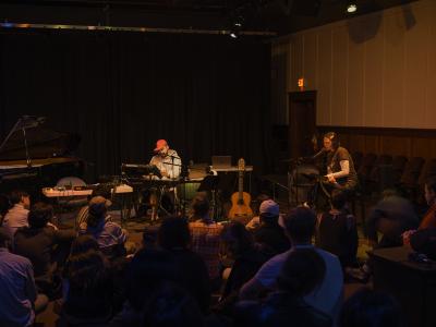 Audience members are seated on the floor of the Grand Luxe Hall facing the performance space where various instruments, microphones, and cables are arranged. Prince Nifty is seated at a synthesizer and wears a pink baseball cap and striped hoodie. On stage right, Kiel Torres is seated on a tall wooden table and reads off of a music stand.
