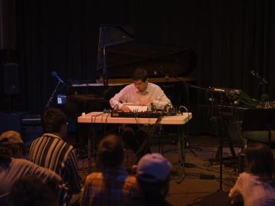 Eddy Wang plays a shruti box while seated at a table next to a synthesizer. A piano, and other instruments and microphones are arranged in the performance space behind him. Eddy is dressed in a light collared shirt. Audience members seated on the floor can be seen from behind.