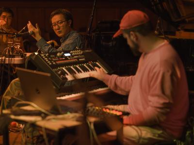 Fan Wu holds an ear of dried corn in his right hand, and holds the back of his left hand against the head of a microphone. Blurred in the foreground of the image, Prince Nifty plays a keyboard. The whole scene is illuminated in a warm orange glow.