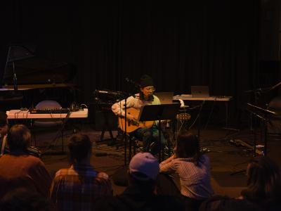 Seated behind a music stand, Julian Hou plays acoustic guitar and sings into a microphone. He wears a black toque, long sleeved shirt, and green pants. A piano,and various electronic instruments and microphones are arranged on the floor and on tables in the background.