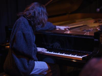 Spencer Cole plays piano with his back facing the camera. His right hand plays the keys while his left hand plucks the strings inside the piano’s frame. He wears a baggy textured sweater over light washed blue jeans.