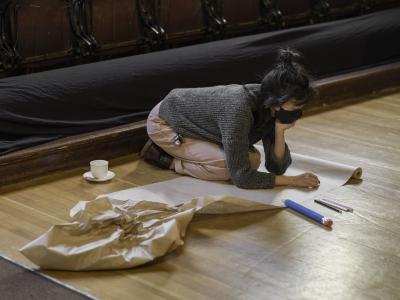 Julia Wong kneels on the wooden floor of the Grand Luxe Hall and writes on a roll of craft paper. They wear a black medical mask, a grey sweater, and pink pants. A teacup, coloured pencil crayons, and a cartoonishly large graphite pencil are placed on the ground next to them.