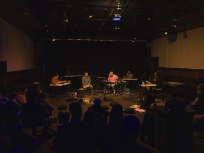 S F Ho kneels on the floor of the Grand Luxe Hall, reading from loose papers and books arranged in front of them. The band is arranged behind them, with Eddy Wang playing synthesizer, Fan Wu seated next to a microphone, Prince Nifty at an electronics station, Julian Hou at a table with a laptop, and Amy Gottung seated with a flute. Meditation pillows, percussive instruments, microphones, and cables are scattered on the floor of the performance space. Audience members can be seen seated from behind.