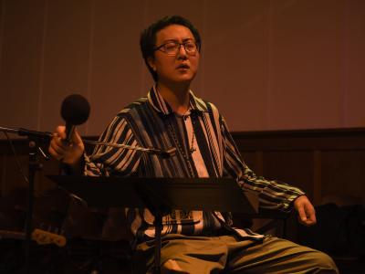 With his eyes closed, Fan Wu sits with his left arm resting on the back of his wooden chair, and his right hand holding a microphone. A music stand is positioned in front of him, and he wears glasses and a beaded necklace over a striped collared shirt. The image has a warm orange glow. 