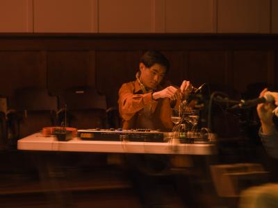 Seated at a table, Eddy Wang dangles interlocking rings into three wine glasses filled with different amounts of water. A microphone points directly into the wine glasses to amplify the sound he’s creating. A synthesizer and transmitter are also positioned on the table. Eddy wears a collared shirt and is bathed in orange light.