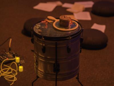 Loose change and small objects are placed on the surface of a tom drum. Meditation cushions and loose papers are scattered on the floor in the background.