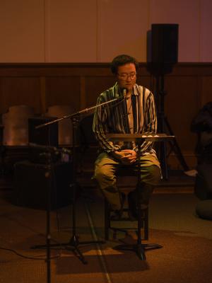 Fan Wu sits on a tall wooden chair and leans into a microphone as he reads off a music stand. He wears glasses and a beaded necklace over a collared shirt with vertical stripes. He’s basked in an orange glow.