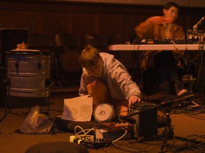 Kasper Feyrer crouches on the floor of the Grand Luxe Hall while adjusting a dial on an amp. Percussive instruments, effects pedals, microphones, bubble wrap, and other sound-making objects are arranged in front of them. In the background, Eddy Wang sits at a table and plays synthesizer.