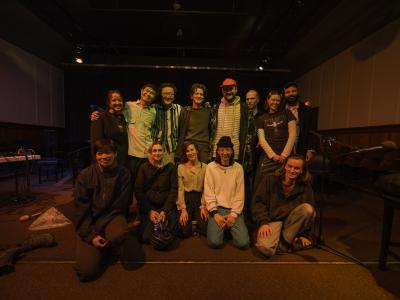 A group photo in the Grand Luxe Hall with performers involved in the Wounded Healers residency. Seven artists stand while five kneel on the ground in front of them.