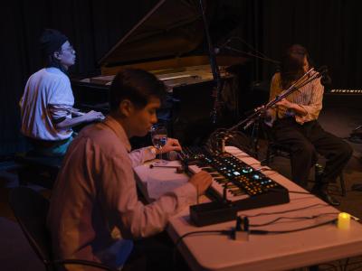 A triangulation of musicians performing in the Grand Luxe Hall. Julian Hou plays piano and is bathed in a blue light. Under an orange glow, Amy Gottung is seated while playing the flute and Eddy Wang is seated at a table while playing synthesizer.