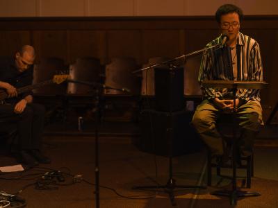 Fan Wu sits on a tall wooden chair and leans into a microphone as he reads from material positioned on a music stand. He wears glasses and a beaded necklace over a striped collared shirt. He’s basked in an orange glow. In the background, Michael Joseph Loncaric sits and plays bass.