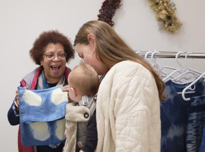 Valérie d. Walker holds an indigo-dyed handkerchief and smiles at a baby that is strapped to a woman wearing a white quilted coat. A rack with Valérie’s other indigo garments can be seen in the background. Valérie has short curly hair and wears glasses and a pink quilted best.
