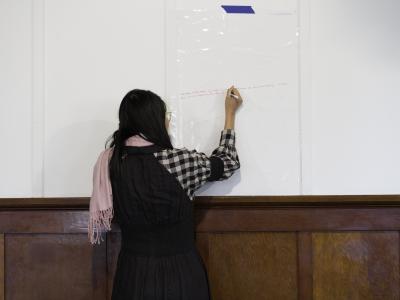 Xinwei Che writes on an erasable surface taped to the wall of the Grand Luxe Hall. Her back is to the camera, and she wears a black and white gingham shirt under a black dress, and a pink scarf.