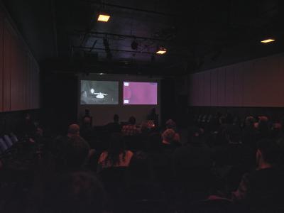 A two-channel video is projected on a screen at the back of the Grand Luxe Hall. The first channel shows a black-and-white image of a birthday candle burning on its side. The second channel shows a chemically manipulated pink film strip. The audience can be seen from behind.
