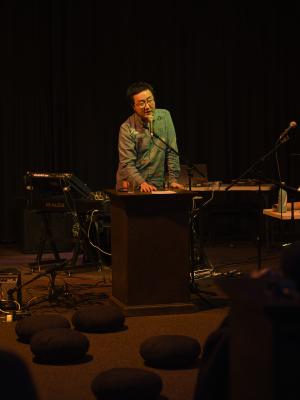 Fan Wu speaks into a microphone from behind a lectern in the Grand Luxe Hall. He is basked in orange overhead light, and wears glasses and a denim shirt. Four black meditation pillows are on the floor in front of him, and various instruments, cables, and microphones can be seen in the background.