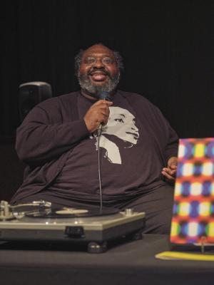 Darius Jones smiles while holding a microphone. He is seated at a table covered with black fabric. A record player and a copy of his LP are placed in front of him. Darius wears glasses and a black cardigan over a black T-shirt printed with a portrait of Sade.