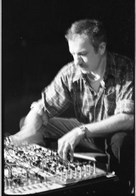 A ¾ medium-long shot of a man looking downward as he adjusts the knobs on a mixer console. One of his hands is blurred. This image is in black and white.