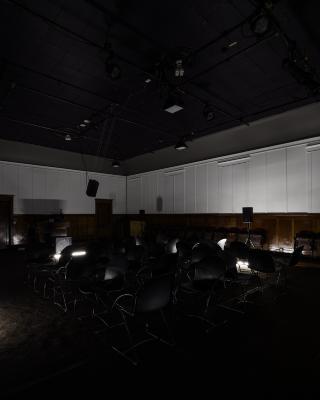 An arrangement of black chairs facing in different directions takes up the centre of the Grand Luxe Hall. The space is dimly lit by tubular lighting fixtures positioned on the floor. A speaker is suspended from the ceiling, and several others are positioned on stands.