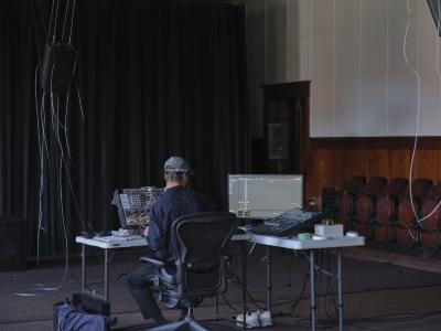 With his back to the camera, Alexi Bari sits in a rolling office chair at a grey folding table in front of a synthesizer patch, computer monitor, and other equipment for composing electronic music. He wears a baseball hat and a long-sleeve button up shirt. A speaker with dangling cables is suspended from the ceiling. 
