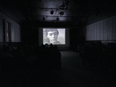 A black-and-white archival video is projected on a screen in the Grand Luxe Hall. The still shows Pascal with her eyes cast down. The space is full of seated audience, who can be seen from behind.  