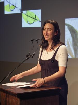Deanna Fong stands at a lectern and moves a set of papers while smiling towards the audience. She wears a white T-shirt under a black knit dress. A microphone is pointed towards her, and an abstract painting is projected on a screen behind her.